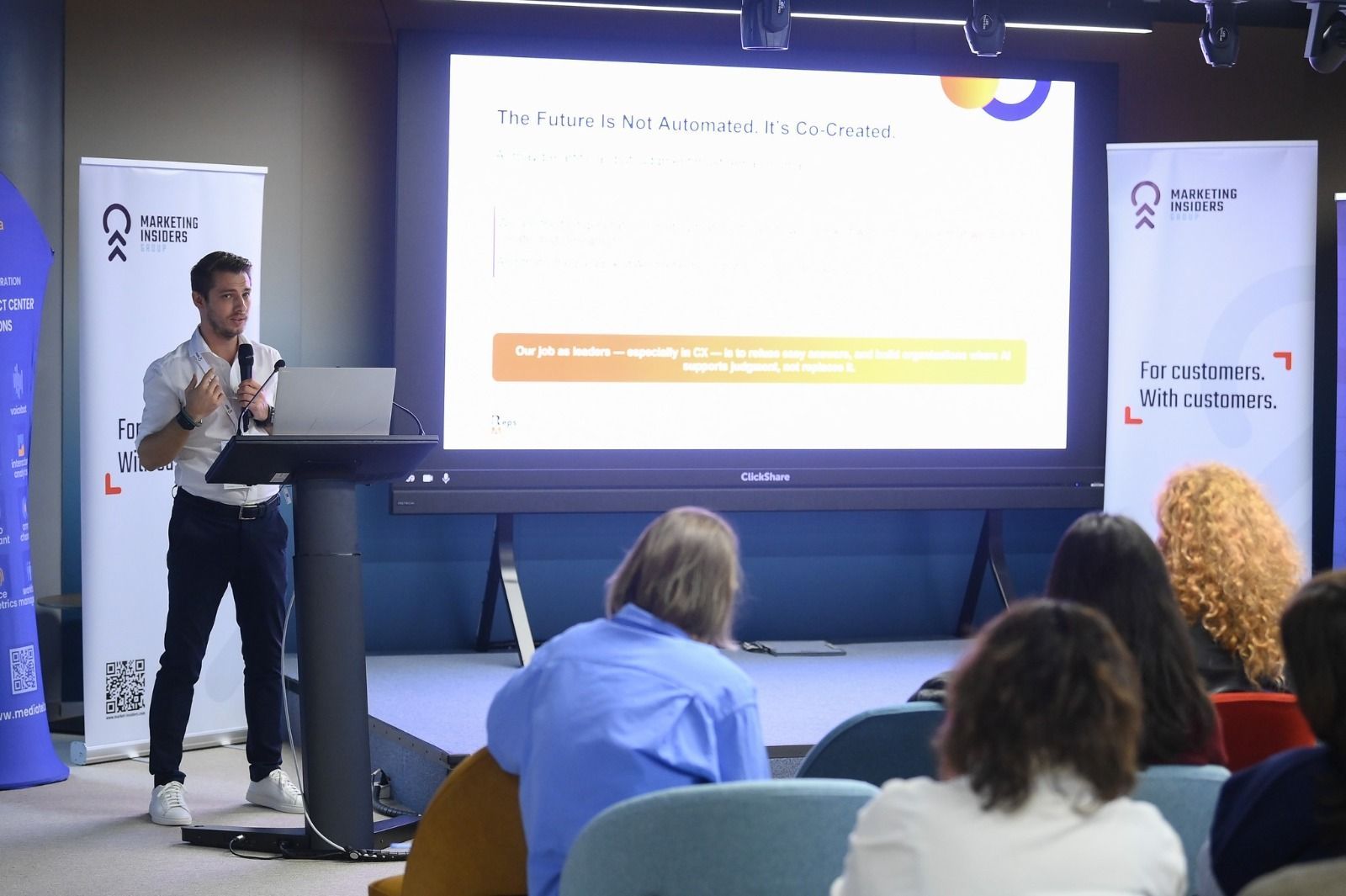 Alin Roșca speaking at a podium beside a slide headed The Future Is Not Automated. It's Co-Created.