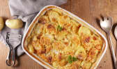 Plat de gratin dauphinois posé sur une table en bois avec pommes de terre, épluche-légume et couverts de service