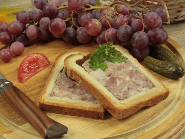 2 tranches de pâté en croûte posées sur un plateau en bois, avec couteau, raisins, tomate et cornichons