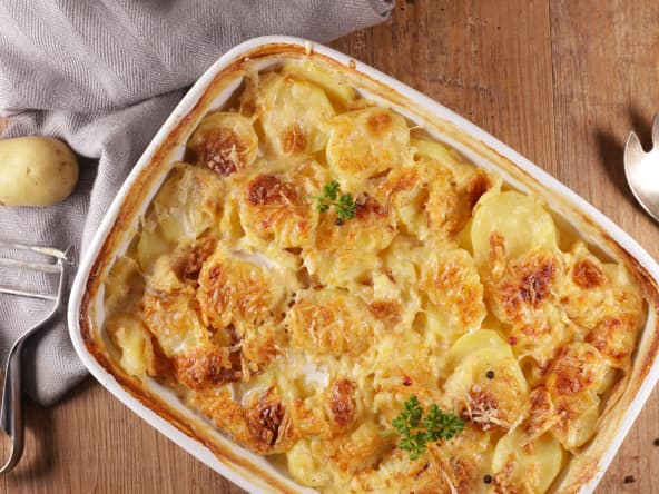 Plat de gratin dauphinois posé sur une table en bois avec pommes de terre, épluche-légume et couverts de service