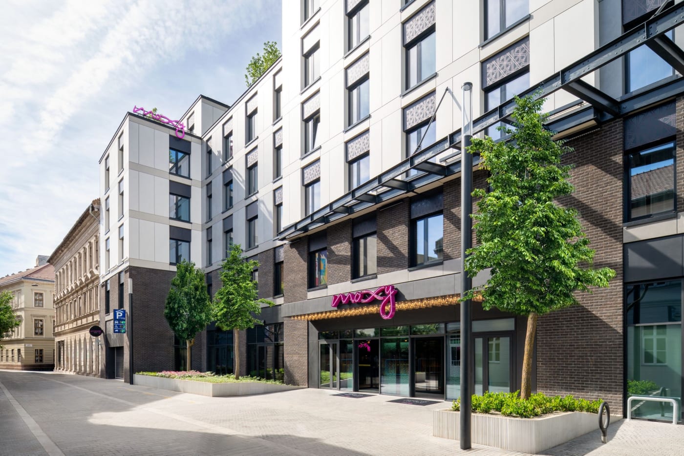 The façade of Moxy Budapest Downtown on Kazinczy Street, with its pink sign and trees out front