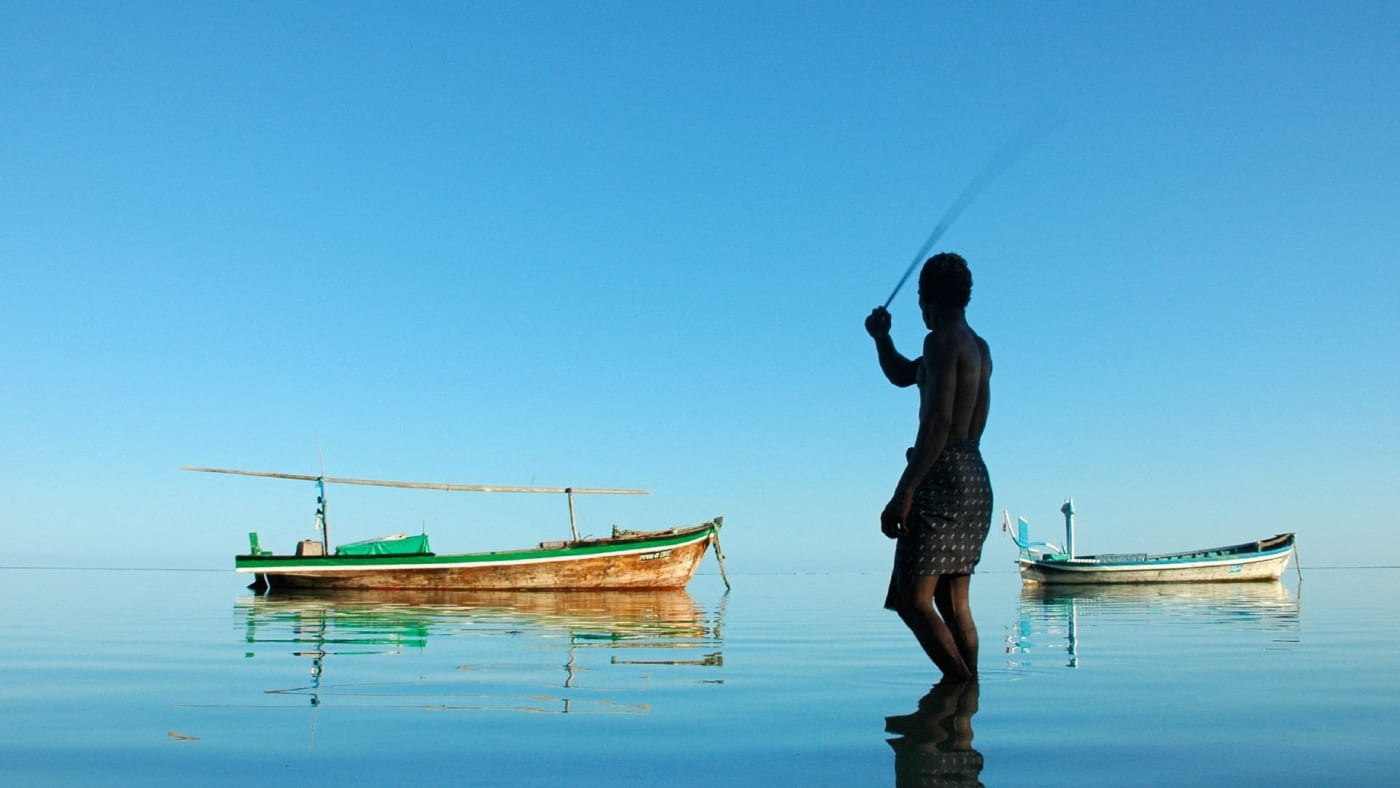 Un pêcheur maldivien lance sa ligne dans un lagon calme, près de deux bateaux traditionnels