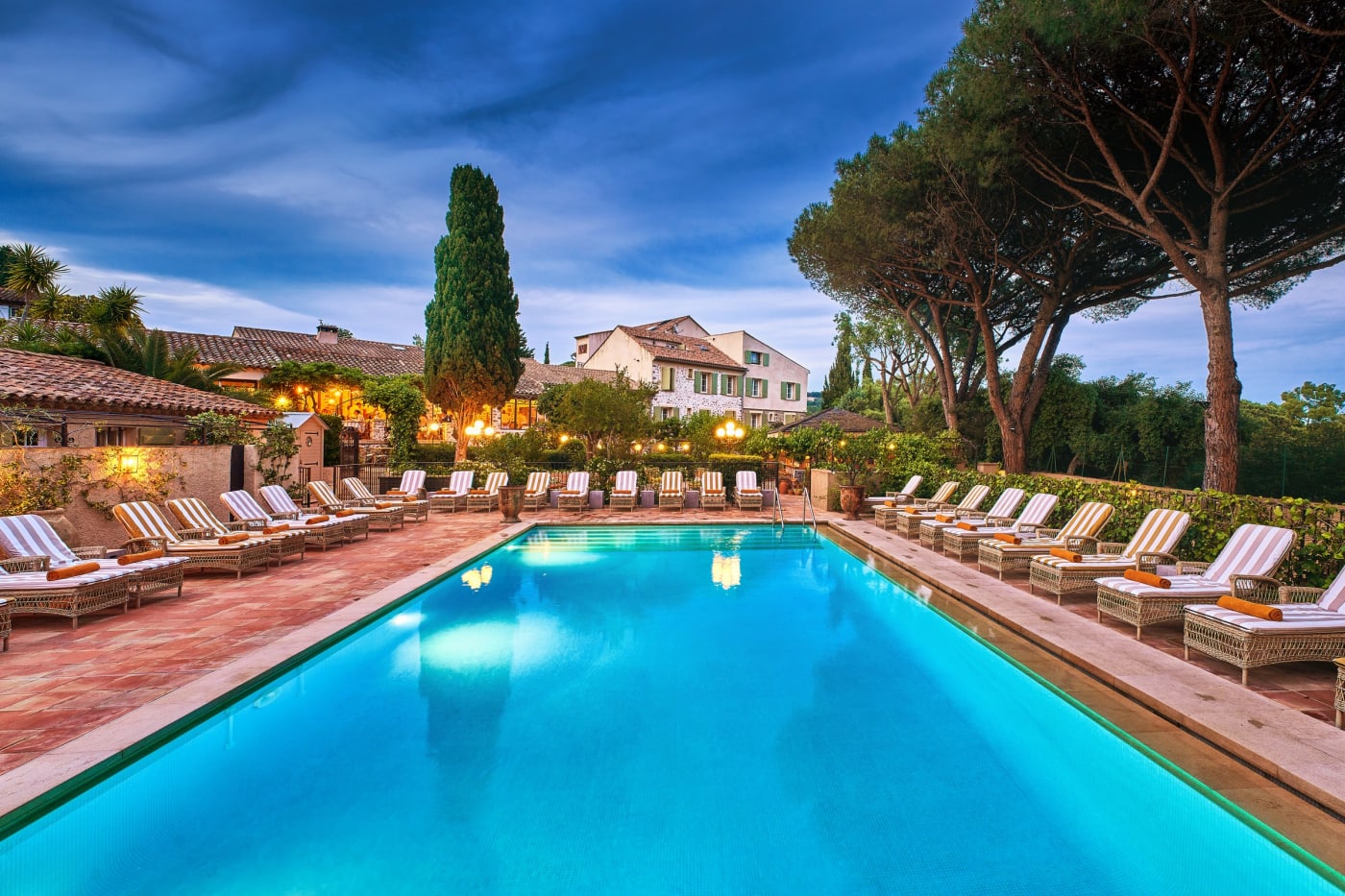 La piscine de La Ferme d'Augustin à Ramatuelle à la tombée du jour, nouveaux transats rayés alignés, pins parasols, cyprès et bâtiments du mas éclairés