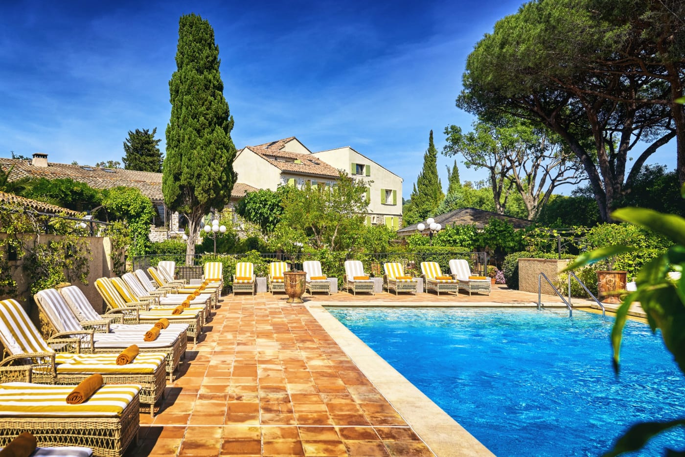 La piscine de La Ferme d'Augustin sous le ciel bleu, rangée de transats rayés jaune et blanc sur les tomettes, cyprès et façades du mas