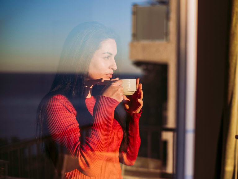 A woman with a coffee looking out of a window at sunrise