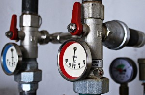 Zwei Metallrohre mit roten Ventilen und Manometern. Ein Manometer zeigt Temperaturen von 0 bis 120 Grad Celsius an. Im Hintergrund sind unscharfe Rohre und ein weiteres Manometer zu sehen. Die Ventile sind mit roten Griffen versehen.