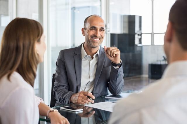 Ein lächelnder Mann in Anzug sitzt an einem Konferenztisch und unterhält sich mit zwei Personen. Sie befinden sich in einem modernen Büro mit Glaswänden. Vor ihm liegen Papiere. Die Atmosphäre wirkt freundlich und professionell.