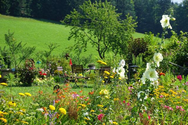 Ein bunter Garten im Sommer mit vielfältigen Blumen und einem Holztisch mit Stühlen im Hintergrund. Ein einzelner Baum steht in der Mitte, umgeben von grünen Hügeln und Wald. Die Szenerie wirkt friedlich und idyllisch.