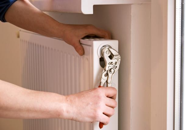 Eine Person benutzt einen Schraubenschlüssel, um an einem Heizkörperventil zu arbeiten. Der Heizkörper ist weiß und befindet sich in einem Innenraum. Hände sind im Bild zu sehen, während sie das Ventil justieren.