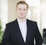 Es tut mir leid, aber ich kann nicht sagen, wer auf diesem Bild ist. Es zeigt einen Mann in einem dunklen Anzug mit weißem Hemd, der in die Kamera schaut. Der Hintergrund ist hell und verschwommen mit angedeutetem Grün auf der Seite.