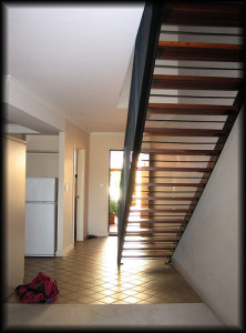 Ein moderner Innenraum mit einer Holztreppe, die schräg nach oben verläuft. Im Hintergrund ist eine Glastür zu sehen, die viel Licht hereinlässt. Links steht ein weißer Kühlschrank und auf dem Boden liegt eine rosa Tasche.