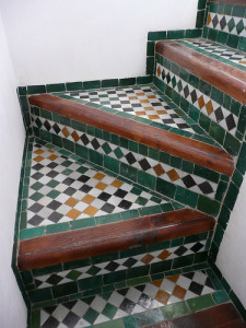 Treppenaufgang aus Keramikfliesen mit geometrischem Muster in Grün, Schwarz, Weiß und Braun. Die Stufen haben holzähnliche Akzente und die Fliesen bilden ein abwechslungsreiches Schachbrettmuster.