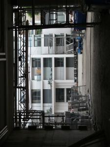 Blick durch ein Baugerüst auf ein modernes weißes Gebäude mit großen Fenstern, von verschiedenen Materialien und Strukturen umgeben. Im Vordergrund sind Bauzäune und ein Betonboden zu sehen.