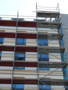 Ein mehrstöckiges Gebäude mit einem Baugerüst an der Fassade. Das Gerüst hat mehrere Plattformen, die mit roten und blauen Schutznetzen gesichert sind. Die Fassade des Gebäudes ist cremefarben mit blauen Fenstern.