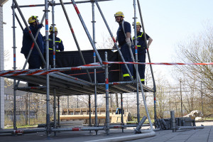 Vier Personen in Arbeitskleidung und Schutzhelmen stehen auf einem Metallgerüst im Freien. Das Gerüst ist mit Absperrband gesichert. Im Hintergrund sind Bäume und ein Maschendrahtzaun zu sehen. Der Himmel ist klar und sonnig.