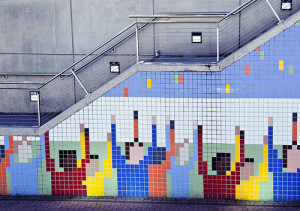 Wandkunst unter einer Treppe, bestehend aus bunten Mosaikfliesen. Die Fliesen bilden stilisierte Menschen in leuchtenden Farben mit erhobenen Armen vor einem blauen Hintergrund, der an einen Himmel oder Horizont erinnert.
