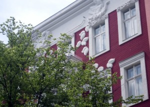 Eine Hausfassade mit roten Ziegeln und dekorativen weißen Stuckelementen an den oberen Fenstern. Im Vordergrund sind grüne Baumzweige zu sehen, die teilweise die Ansicht des Gebäudes verdecken.