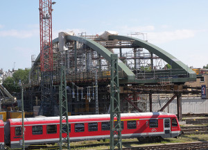 Eine im Bau befindliche Brücke mit einem großen Bogen aus grünem Stahl wird von Gerüsten und Kränen umgeben. Unterhalb der Brücke fährt ein roter Zug auf den Gleisen entlang. Im Hintergrund ist ein blauer Himmel zu sehen.