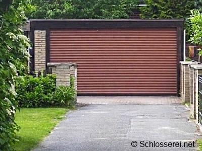 Fertige Garage nach Abschluss der Bauarbeiten Braune Garage mit Rolltor und umliegendem Garten. Der Eingang der Einfahrt ist mit einem niedrigen Mauerwerk auf der linken Seite flankiert. Umgeben von grüner Vegetation, vermittelt das Bild ein ruhiges, ländliches Ambiente.