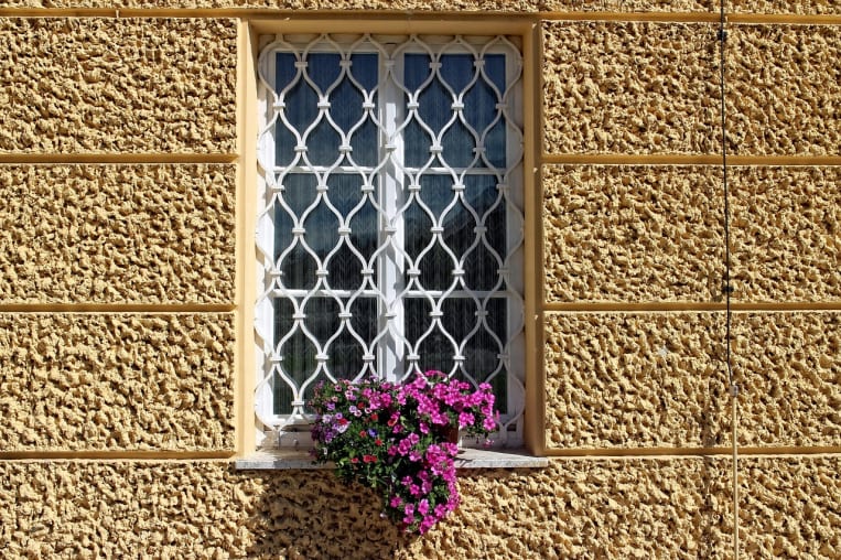 Ein weißes Fenster mit dekorativem Gitter, das in eine beige, strukturierte Wand eingelassen ist. Vor dem Fenster steht ein Blumenkasten mit lila und rosa Blüten, die lebendig hervorstechen.