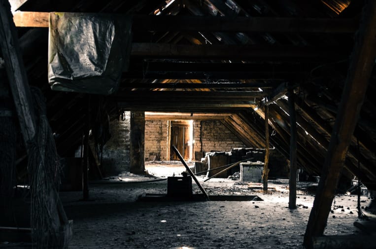 Ein dunkler, verlassener Dachboden mit Holzbalken und einem staubigen Boden. Ein schwaches Licht fällt durch offene Türen in der Ferne und beleuchtet teilweise den Raum. Verschiedene Gegenstände und Schutt sind verstreut.