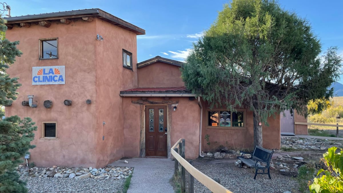 A rustic adobe-style building with a sign reading 'La Clinica' on the front wall. The clinic has natural, earthy tones, blending with the surrounding landscape. A tree and small shrubs are situated near the entrance, which is accessible by a pathway with a handrail leading to a wooden front door. The peaceful setting and natural materials create a welcoming atmosphere.