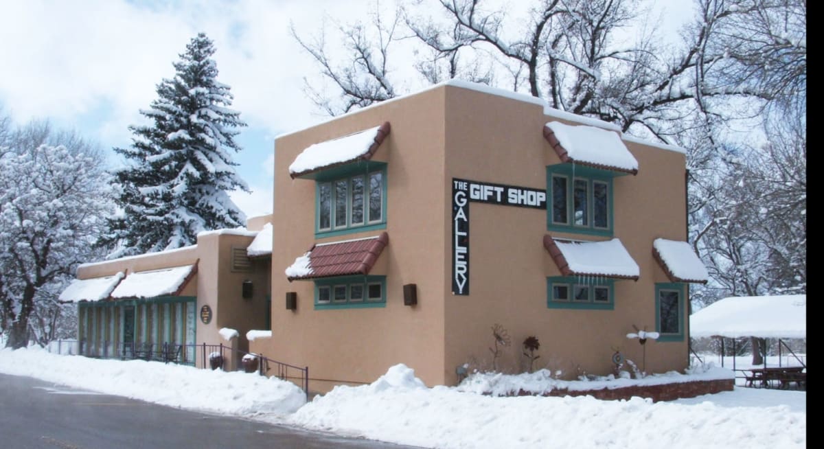 Photo of a stucco building with a sign reading 'The Gallery Gift Shop' and the address number '132' mounted on the corner. The building features turquoise window frames and red tile awnings, with a small garden of lavender and ornamental plants in the foreground. The sky is blue with scattered clouds.