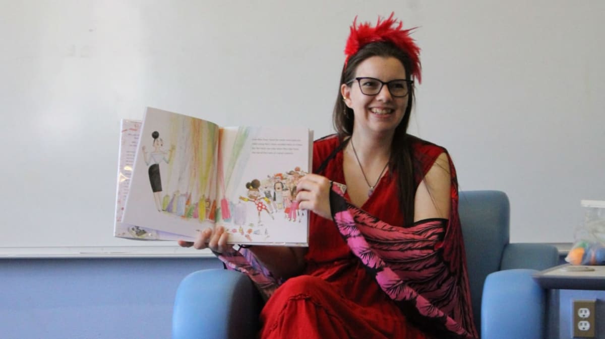 A woman dressed in bright colors and wearing large feathered wings holds up a children’s book while reading to a group of young kids. She is seated in a library, surrounded by colorful props and story-themed decorations, with bookshelves in the background.