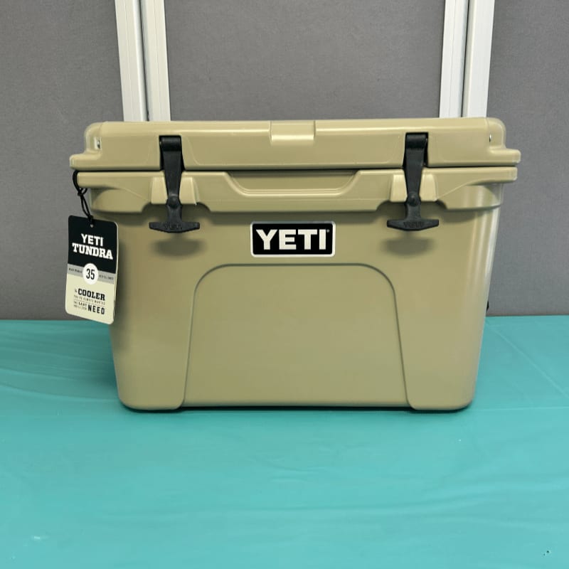 A tan YETI cooler with a tag sits on a teal surface in front of a white frame.