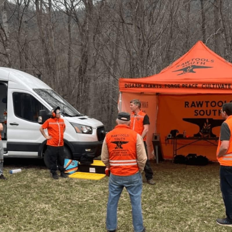In the foreground, several people in orange vests stand near a white van and an orange tent, while the background shows a wooded hillside.