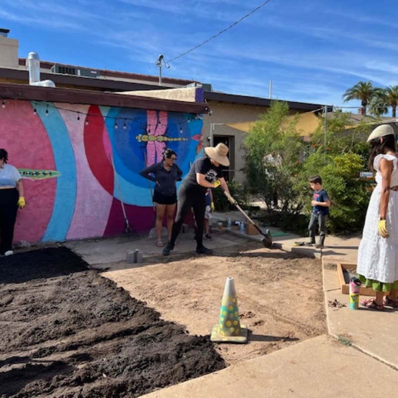 In the foreground, people are working on a garden bed with dark soil, while in the background, a colorful mural adorns a building under a clear blue sky.