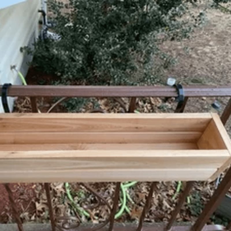 A new wooden planter box is attached to a brown railing in front of a house with green foliage.