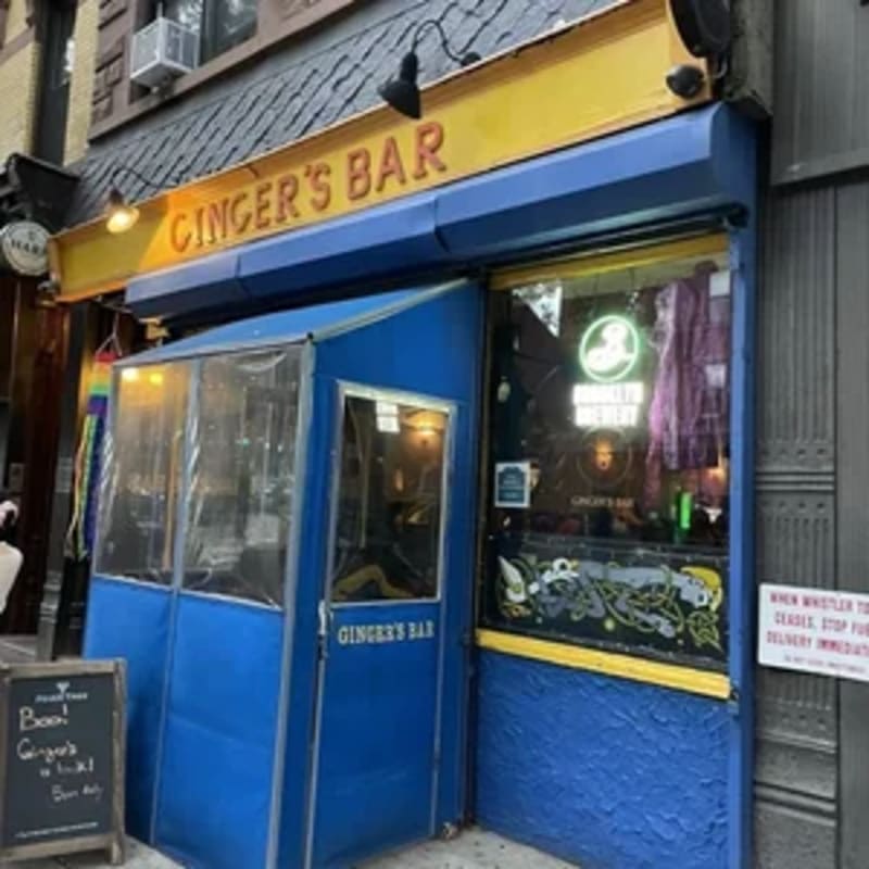A blue enclosed entryway with a sign for "Ginger's Bar" stands in front of the bar's storefront, with a chalkboard sign to the left and a brick building in the background.