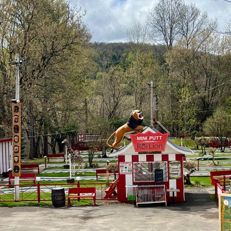 A miniature golf course with a roaring lion statue in the foreground is set against a backdrop of lush green trees and a cloudy sky.