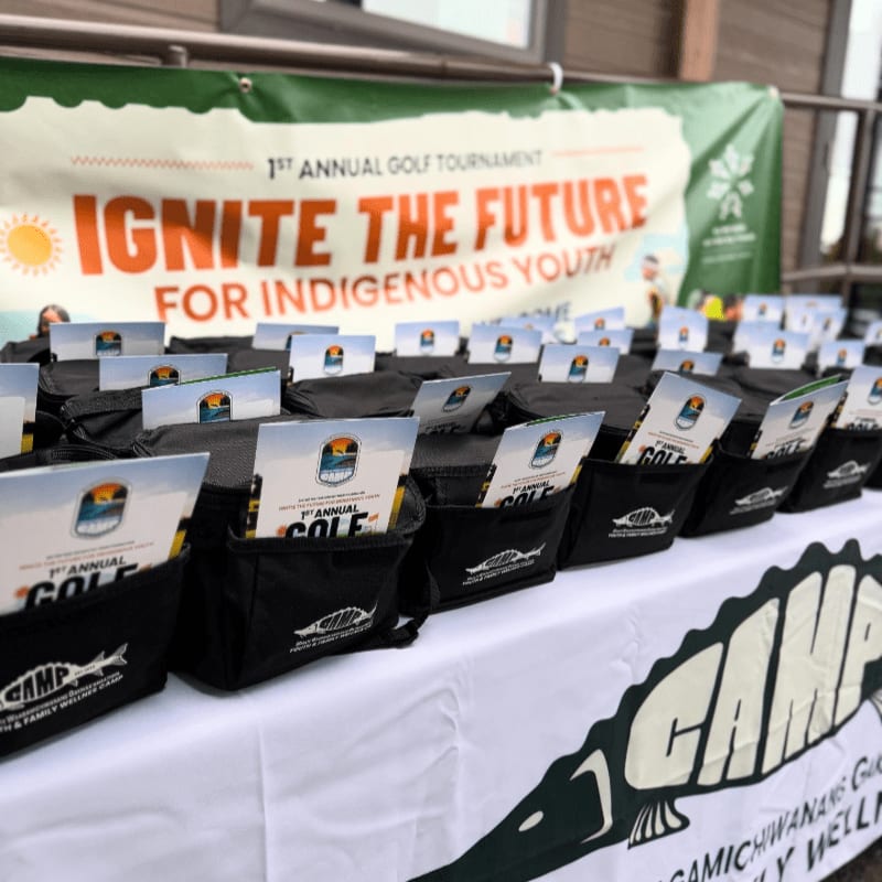 Rows of black gift bags filled with brochures and other items sit on a table in front of a banner advertising the "1st Annual Golf Tournament Ignite the Future for Indigenous Youth."