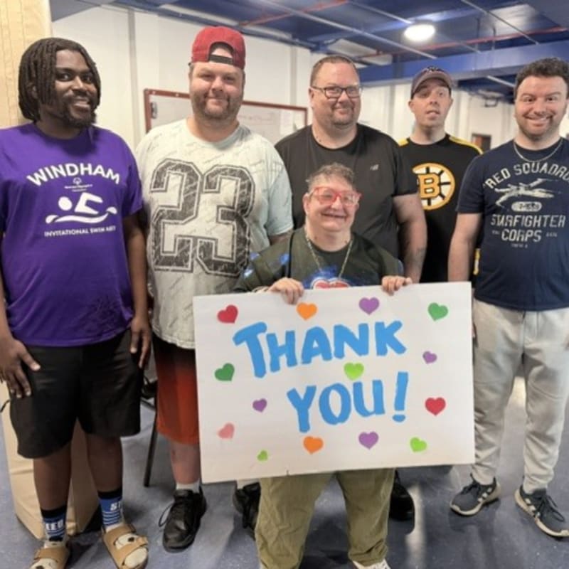 Six people stand together, with one in the foreground holding a "Thank You!" sign, against a backdrop of a room with a whiteboard and sports equipment.