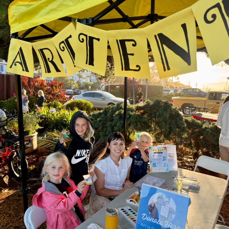 Children and an adult smile behind a table with art supplies under a yellow tent with "ART IS FUN" banners, while a street with parked cars and trees forms the background.
