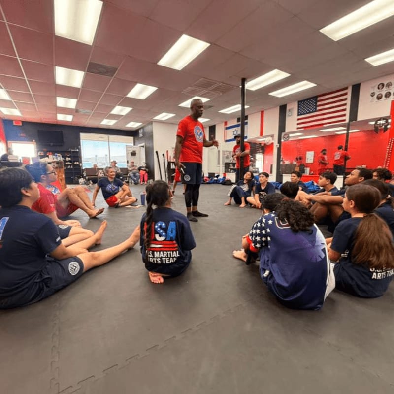 A group of children in a martial arts class sit on the floor in the foreground, listening to an instructor in a red shirt in the background, with American flags and training equipment visible.