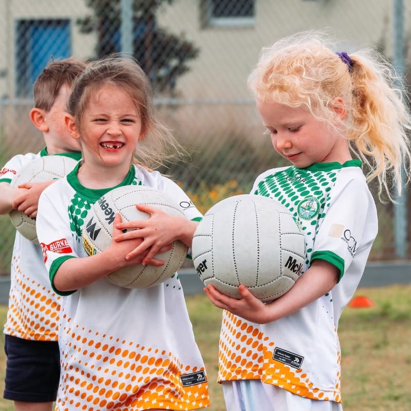 Two young girls in matching jerseys hold Gaelic footballs in the foreground, with a blurred background of a fence and building.