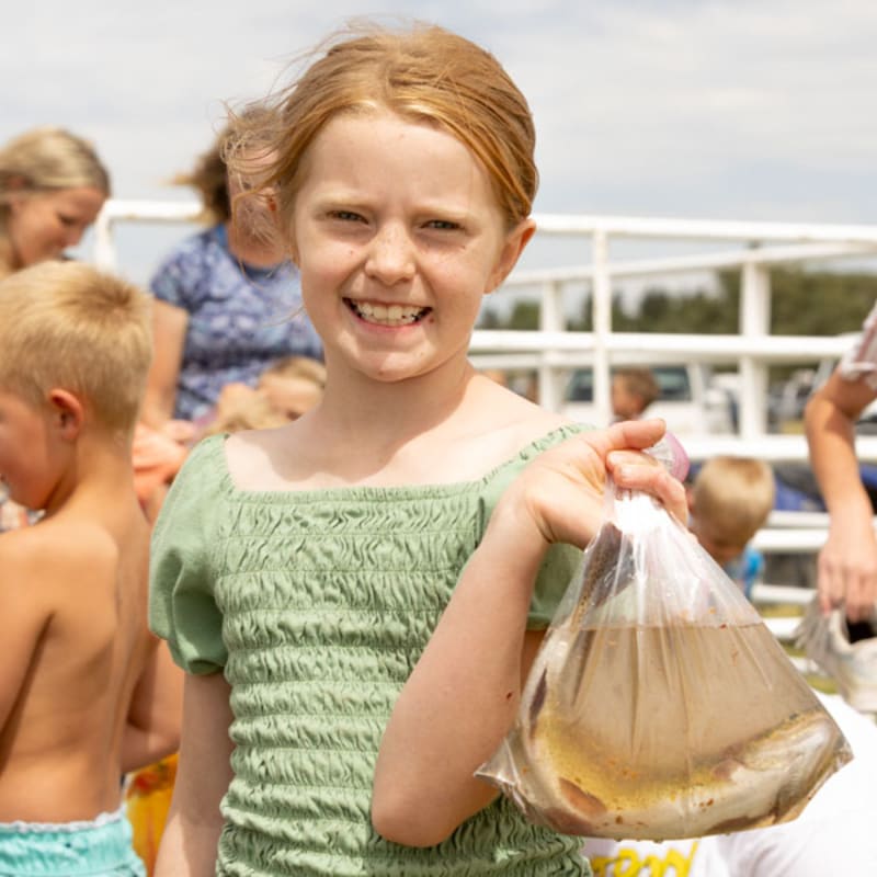 A smiling young girl with red hair holds up a plastic bag filled with fish in the foreground, while other children and adults are blurred in the background under a bright sky.