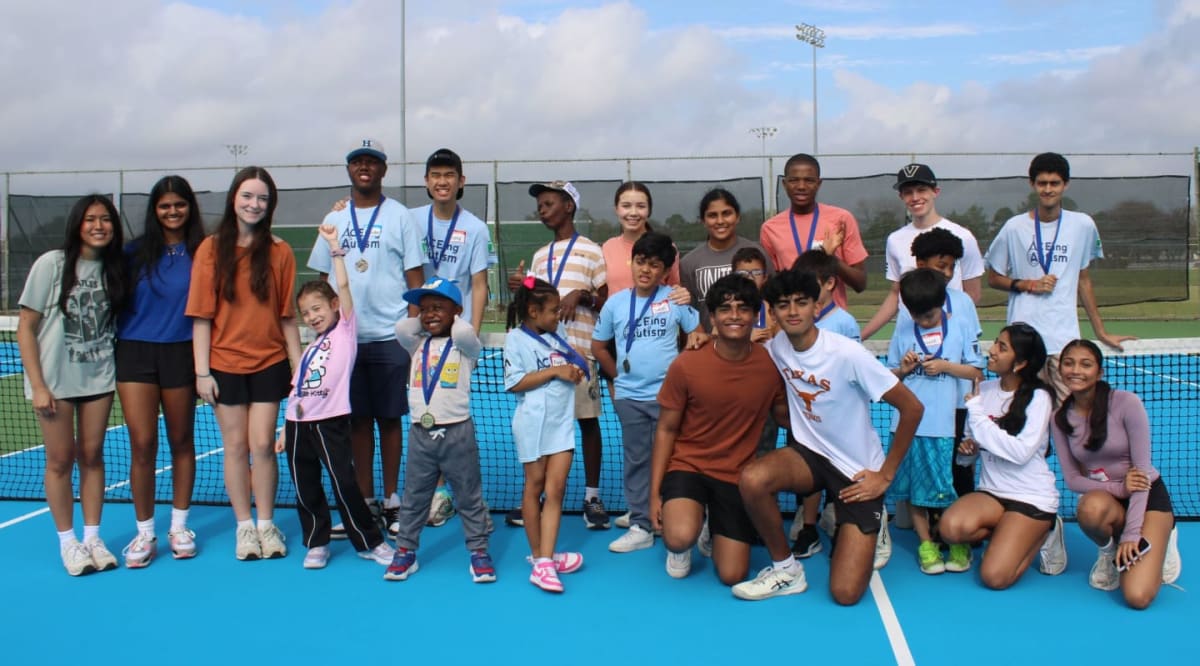 A group of children and young adults pose for a photo on a blue tennis court with a net in the foreground and a cloudy sky with distant trees and lights in the background.