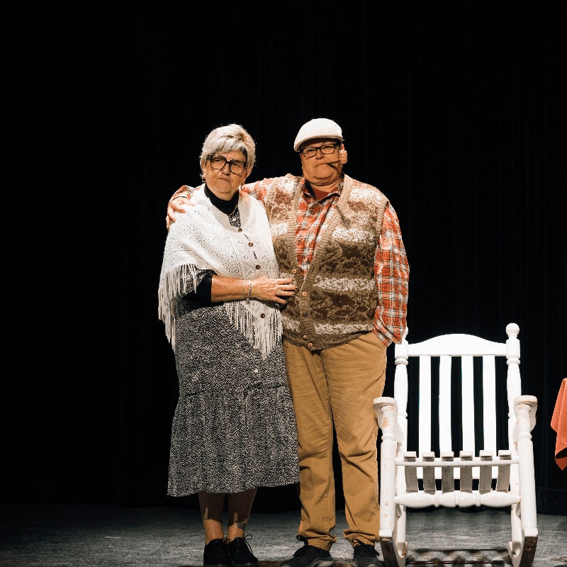 A couple stands on a stage with a rocking chair in the foreground and a black curtain in the background.