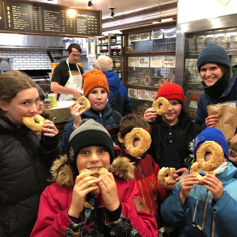 A group of children in winter coats and hats are happily eating bagels in the foreground, while a baker works in the background of a bustling bagel shop.