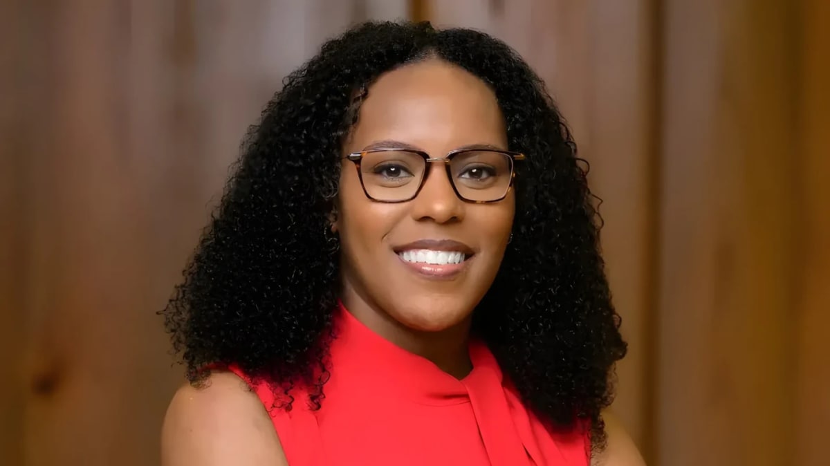 A smiling woman with glasses and curly hair in a red top is set against a blurred wooden background.