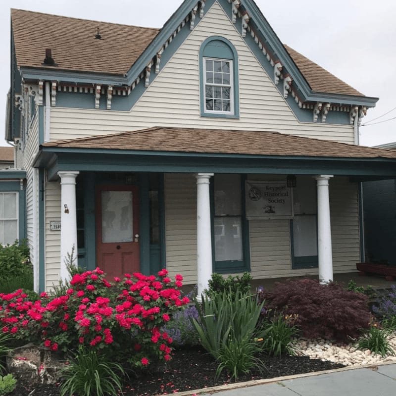 Vibrant pink flowers bloom in the foreground before a charming, light-colored Victorian house with a welcoming porch.