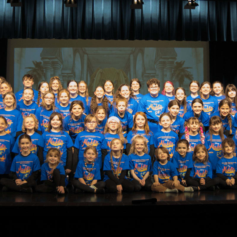 A large group of children in matching blue shirts stand on a stage in front of a projected backdrop of an ornate building.