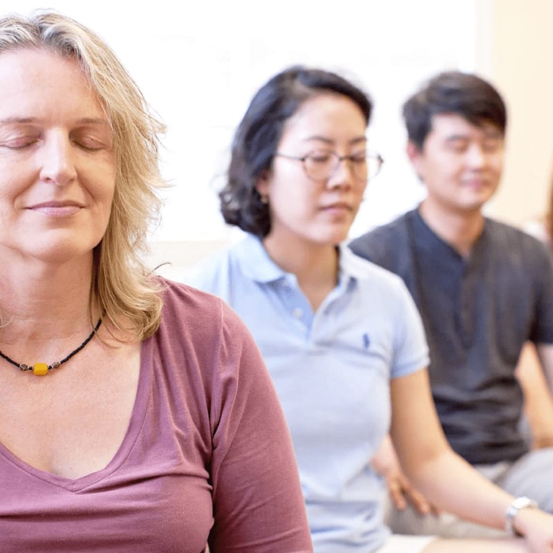 A woman with blonde hair and a purple shirt meditates in the foreground, while people of Asian descent with their eyes closed sit behind her in a softly lit room.