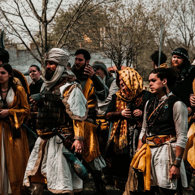 A group of people in historical costumes stand in a grassy area with trees in the background.