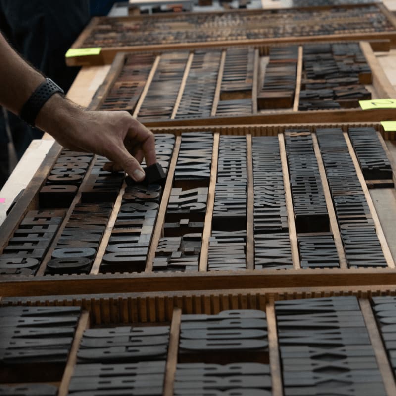 A hand reaches into a wooden tray filled with antique metal letterpress type, with more trays of type visible in the blurred background.