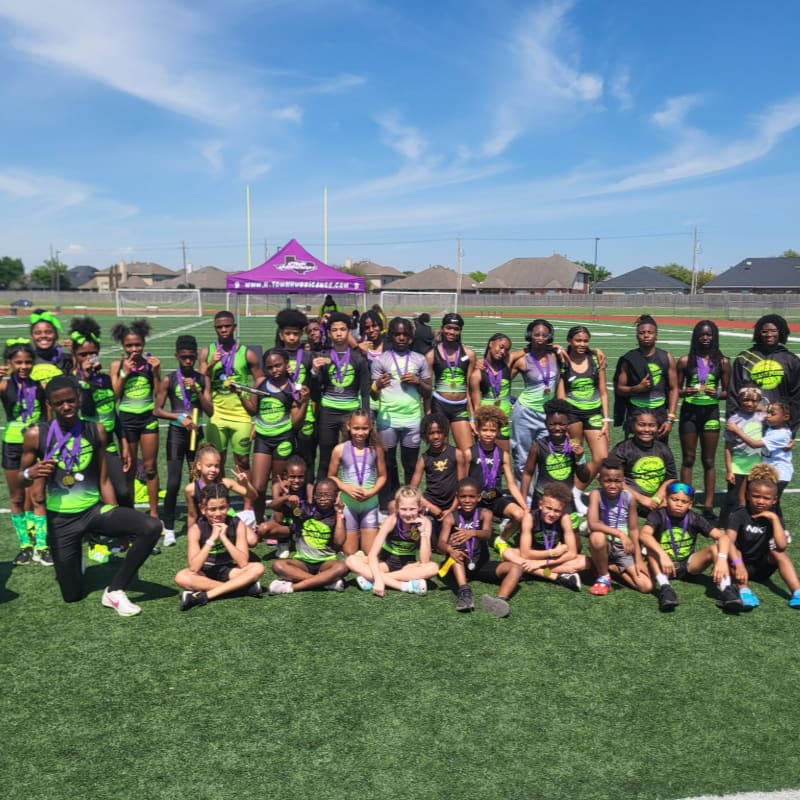 A large group of young athletes in neon green and black uniforms pose on a green turf field with a purple banner and goalposts in the background under a bright blue sky.